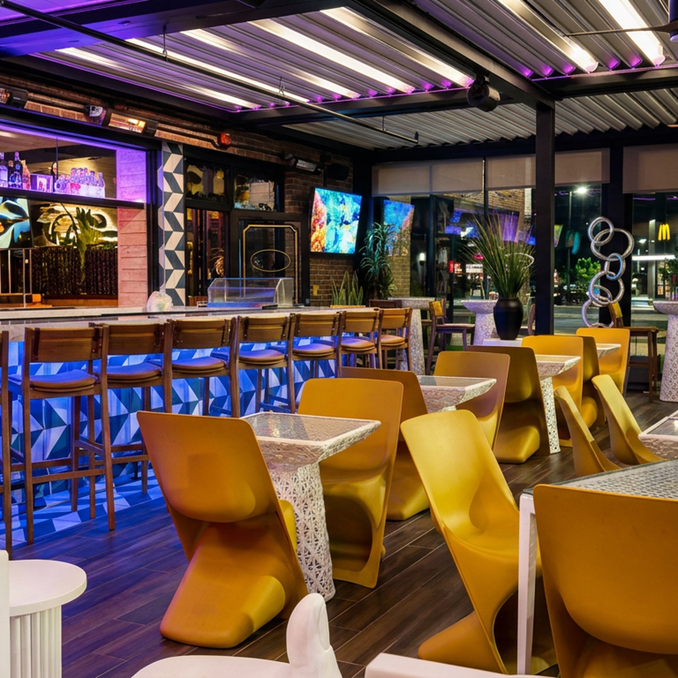 Vibrant restaurant interior with yellow chairs, mosaic tables, blue-purple lighting, bar seating, plants, and wall-mounted screens.