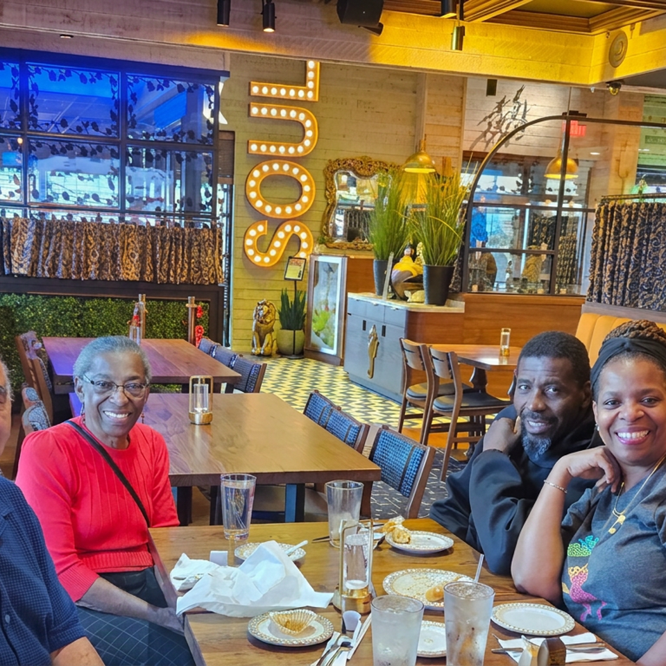 Four diners smile together at a restaurant table surrounded by colorful decor, patterned curtains, plants, and warm lighting.
