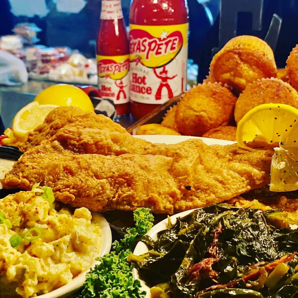 Platter of fried seafood, hush puppies, macaroni salad, greens, lemon wedges, dipping sauce, and hot sauce bottles.